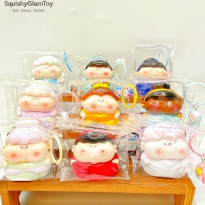 May include: A collection of SquishyGlamToy keychains, each featuring a cartoon character with a round face and various hair colors and outfits. The keychains are packaged in clear plastic and displayed on a wooden shelf. The brand's logo is visible at the top.