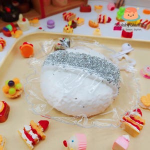 以下が含まれることがあります： 銀色のグリッターバンドが付いた、白くテクスチャ加工された丸い物体で、透明なプラスチックで包まれ、ミニチュアのカラフルな食品玩具に囲まれています。画像には「Squishy GlamToy: Soft. Sweet. Stylish.」というテキストが含まれています。