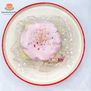 May include: A pink, round squishy toy with colourful sprinkles, presented on a white plate with a red rim. The toy is wrapped in clear plastic and sits on a yellow base. The plate has a decorative lace doily.
