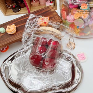 Op de afbeelding: Een doorzichtige plastic zak gevuld met rode, gelei-achtige snoepjes, op een zilveren dienblad. Andere miniatuursnoepjes, waaronder macarons en koekjes, zijn zichtbaar. Een pot vol kleurrijke miniatuursnoepjes staat op de achtergrond.