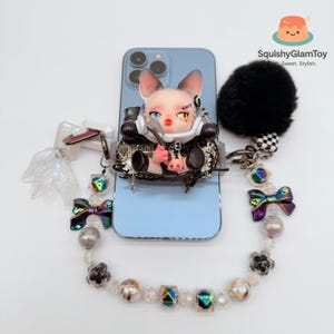 May include: A light blue phone case with a distinctive design. It features a doll-like figure holding a spoon. The case includes a black pom-pom and a beaded strap with iridescent bows and beads. The doll has the word "Guilty" on its face.