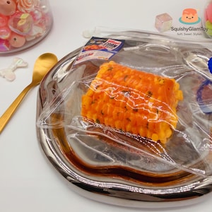 Puede incluir: Un juguete blando realista de mazorca de maíz naranja y amarillo, empaquetado en una bolsa de plástico transparente, se encuentra en una bandeja plateada. Se ve una pequeña caja de cereales. Una cuchara dorada y objetos decorativos están en el fondo.