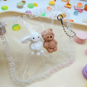 May include: A clear, square pouch with star and lace trim, containing a white bunny and a brown teddy bear charm. The pouch has a silver chain and ring attachment. Miniature sweets are scattered around the pouch.
