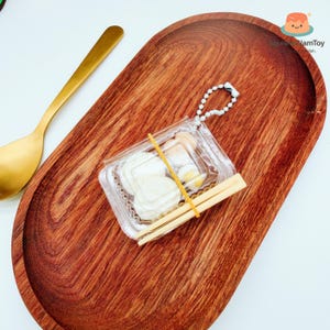 May include: A miniature food set in a clear plastic container, secured with a rubber band, and includes two wooden chopsticks. The set is on a wooden oval tray with a gold spoon. The tray has a rich, reddish-brown colour.
