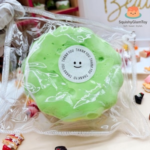 May include: A green, squishy toy in a clear plastic bag. A white sticker with a smiling face and the words "Thank You" is on the toy. The brand name "SquishyGlamToy" is visible in the background.