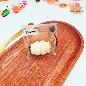 May include: A clear plastic bag containing a cream-coloured, squishy pastry with a brown filling. The bag has the word "Every" on a white label. The pastry is attached to a black beaded chain. Other miniature food items are in the background.