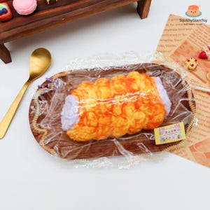 May include: A faux pastry, resembling a cream horn, is presented on a wooden tray, wrapped in clear plastic. The pastry has a golden-brown exterior and a white, creamy filling. A gold spoon and other miniature desserts are also visible.