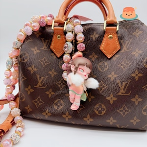 以下が含まれることがあります： 茶色のモノグラムハンドバッグ、茶色の革のディテール。ピンクと白のトーンの装飾的なビーズストラップがバッグからぶら下がっています。ピンクの衣装と白い毛皮のトリムが付いた小さな人形のチャームが取り付けられています。