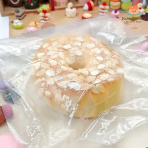 May include: A close-up of a squishy toy donut with a hole in the center, covered in almond slices. The donut is wrapped in clear plastic. The background features other miniature toy desserts.