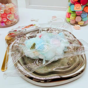 Peut inclure: Un sac en plastique transparent contenant un bouquet de fleurs pastel en tissu, posé sur une assiette argentée à bords festonnés. Des bocaux de perles et de macarons sont en arrière-plan.