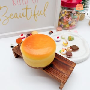 May include: A round, orange-topped cake sits on a small wooden stand. The cake has a light yellow base and a darker orange top. The background includes a framed sign with gold lettering that reads "Kind of Beautiful."