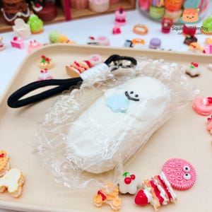 May include: A white, cloud-shaped squishy toy with a blue star and a smiling face, attached to a black lanyard. The toy is packaged in clear plastic with a lace trim. Miniature food items surround the toy.