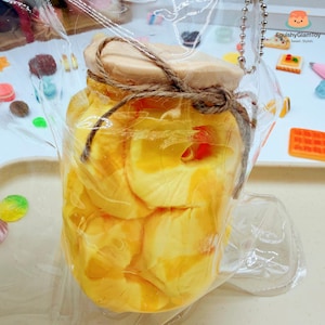 May include: A jar filled with yellow petals, sealed with a wooden lid and twine, encased in clear plastic. The jar is hanging from a silver chain. The text "SquishyGlamToy" is visible in the background.
