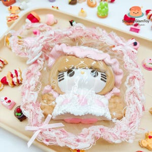 May include: A kawaii-style doll with a light brown face, wearing a pink bonnet and white dress, is encased in a pink lace frame. The doll is surrounded by miniature desserts on a beige tray, creating a whimsical, sweet theme.