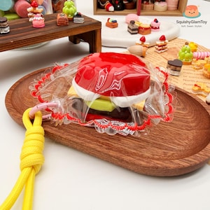Pode incluir: Um brinquedo de hambúrguer em miniatura vermelho, branco, amarelo e castanho está envolto em plástico transparente com uma borda de renda vermelha. O hambúrguer está em uma bandeja oval de madeira, com um cordão amarelo preso.
