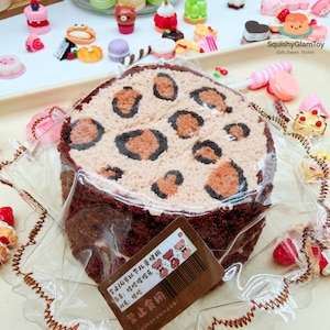May include: A round, brown cake with a tan top decorated with brown leopard spots. The cake is wrapped in clear plastic and surrounded by miniature, colorful desserts. A small brown tag with text and a barcode is attached to the cake.