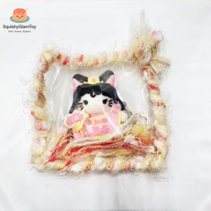 Pode incluir: Figura de gato estilo kawaii com roupa rosa e amarela, cabelo preto e coroa, dentro de um quadrado de plástico transparente. A moldura em volta da figura é feita de fio fofo e multicolorido. O logotipo "SquishyGlamToy" é visível.