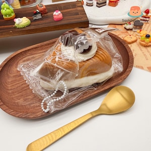 Puede incluir: Un juguete blando con forma de pastel en un envoltorio de plástico transparente, sobre una bandeja ovalada de madera. El pastel tiene una combinación de colores marrón, blanco y crema, con una cadena de cuentas blancas. Una cuchara dorada en primer plano.