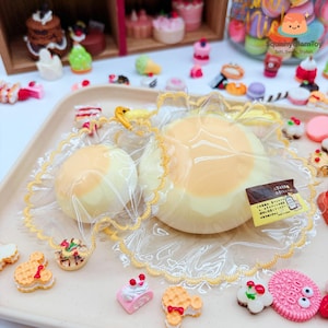May include: A large, round, yellow and white squishy toy, packaged in clear plastic with a yellow lace trim. The toy is surrounded by miniature food items, including cakes, cookies, and macarons. The text "Squishy GlamToy" is visible.