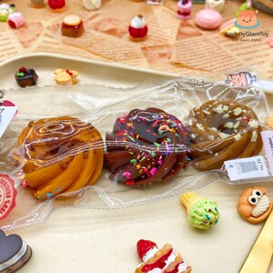 May include: A collection of miniature food items, including a swirl pastry, a chocolate dessert with sprinkles, and a caramel pastry. Other items include a miniature ice cream cone and a small cake. The items are in a clear plastic bag.