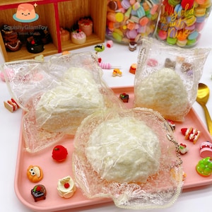 May include: A collection of miniature, cream-colored, squishy toys in clear plastic packaging, arranged on a pink tray. The scene includes various small, colorful dessert-themed charms and a gold spoon, suggesting a playful, collectible theme.