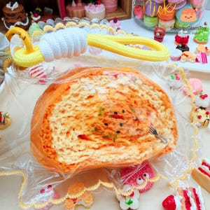 May include: A large, orange and white bread-shaped squishy toy, wrapped in clear plastic. The toy has a yellow and white handle. The background includes other miniature food toys.