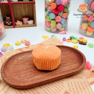 Op de afbeelding: Een oranje cupcake met witte spikkels ligt op een houten ovaal dienblad. Potten gevuld met kleurrijke macarons en miniatuur taartjes staan op de achtergrond. De afbeelding bevat de tekst "Squishy Glam Toy: Soft, Sweet, Stylish."