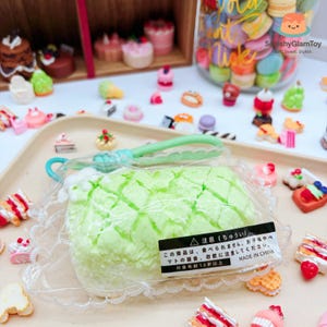 May include: A green, squishy toy shaped like a melon bun, packaged in clear plastic. The image features various miniature dessert toys, including cakes and pastries, and a jar filled with colorful macarons. The text on the package warns against consumption.