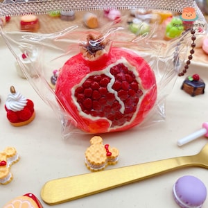 Op de afbeelding: Een felrode granaatappel, gedeeltelijk doorgesneden om de sappige, donkerrode zaden te onthullen, is het middelpunt. De afbeelding bevat miniatuurvoedsel zoals cakes en koekjes, en een goudkleurige lepel. Het thema is voedsel.