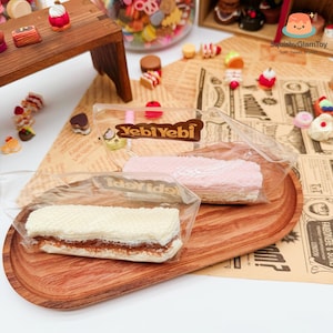 May include: A wooden serving tray holds two packaged wafer cookies, one white and one pink. The white wafer has a chocolate filling, while the pink one appears to be strawberry flavored. The packaging reads "Yebi Yebi". Miniature desserts are scattered around.