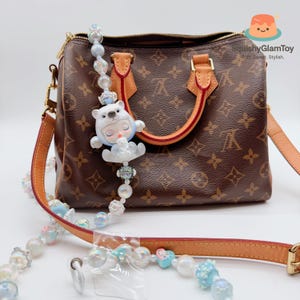 May include: A brown handbag with a monogram pattern, featuring a decorative beaded strap with a sleeping bear charm. The strap includes white, blue, and iridescent beads. The bag has tan leather handles and trim.