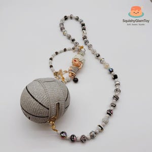 May include: A silver-colored, spherical coin purse with black stripes and a gold zipper, attached to a beaded necklace. The necklace features a small figurine and various beads in black, white, and clear colors. The brand name "SquishyGlamToy" is visible.