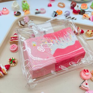 Peut inclure: Un objet rectangulaire rose et blanc avec des paillettes est enfermé dans du plastique transparent. L'objet est entouré de jouets miniatures sur le thème de la nourriture, notamment des gâteaux, des biscuits et des pâtisseries, sur un plateau de couleur claire.