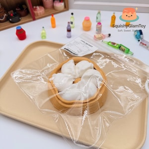 May include: A miniature dim sum set featuring white dumplings in a bamboo steamer, encased in clear plastic. The set is on a beige tray, with miniature bottles and desserts in the background. The text "SquishyGlamToy" is visible.