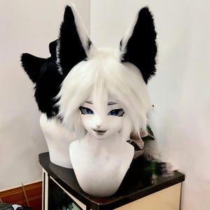 May include: A white and black furry cosplay head with blue eyes and a smiling expression. The head has large, upright ears with black tips and white fur. Another similar head is visible in the background.