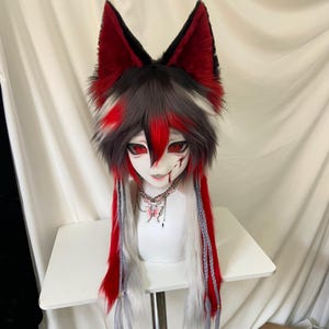 May include: A full-head mask featuring tall, pointed, red and black faux fur ears, layered grey and red hair, and a white face with red eyes and a red mouth marking. The mask includes long, flowing red and white hair strands with braided accents.