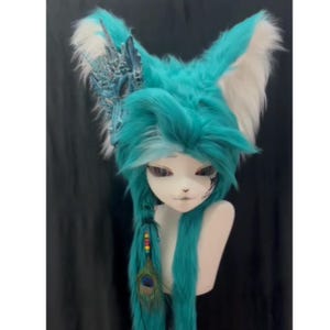May include: A teal and white furry fox ear headpiece with long teal hair and a decorative blue dragon-like accent. The wig features a braid with colorful beads and a peacock feather. The headpiece is displayed on a mannequin head.