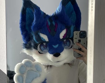 May include: A blue and white furry wolf head fursuit with large cartoon eyes and a paw raised in a wave. The fursuit has blue and white fur with accents of purple and pink. The fursuit is being reflected in a mirror.