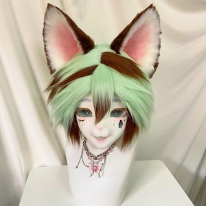 May include: A white mannequin head with a furry, green and brown wig and large, pointed ears. The face has detailed eye makeup and small facial tattoos. A silver chain necklace with a pink butterfly pendant completes the look.