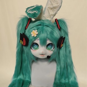 May include: A teal-colored cosplay costume featuring a large, expressive face with oversized eyes and a small microphone. The costume includes long teal hair, bunny ears adorned with pearls, and a flower accent. The overall design is whimsical and detailed.