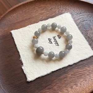 May include: A beaded bracelet featuring gray and white round stones, interspersed with smaller white beads. The bracelet rests on a small, off-white cloth with the words "oh happy day" printed on it. A gold-colored accent is visible.