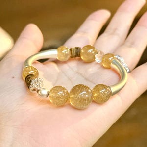 May include: A bracelet featuring translucent golden rutilated quartz beads, smooth silver-toned curved metal segments, small dark brown wooden spacers, and a silver-toned decorative bead with a textured surface and clear crystals.