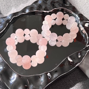 May include: Two rose quartz bead bracelets, each with varying sizes of round beads. The bracelets are a soft pink color and are displayed on a reflective, silver-toned tray. The beads have a smooth, polished appearance.