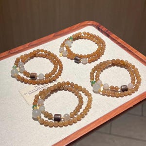 Op de afbeelding: Vier amberkleurige armbanden met kralen worden gepresenteerd op een houten dienblad. Elke armband heeft kleine, ronde kralen, afgewisseld met lichtere kralen, kleine groene kralen en donkerbruine cilindrische kralen. De armbanden zijn in een cirkelvorm gerangschikt.