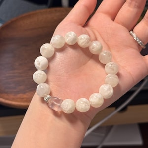 Könnte beinhalten: Ein weißes Perlenarmband mit großen, runden Perlen. Das Armband hat eine kleine, klare, rechteckige Perle und eine silberfarbene Metallperle. Das Armband wird auf der Hand einer Person präsentiert.