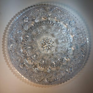 Op de afbeelding: Transparante glazen serveerschaal met ingewikkelde bloemen- en sterpatronen. De schaal heeft een gedetailleerd, gestructureerd ontwerp met een geschulpte rand. Het midden van de schaal heeft een stervormig ontwerp.