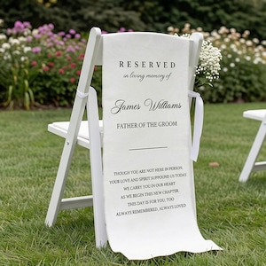 May include: White wooden chair with a fabric banner that reads "RESERVED in loving memory of James Williams FATHER OF THE GROOM." The banner also includes a heartfelt message. The chair is decorated with white flowers and ribbon.
