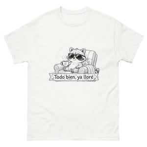 May include: A white t-shirt featuring a black and white line art graphic of a raccoon wearing sunglasses, sitting in an armchair while holding a coffee mug. Below the raccoon is a banner with the text "Todo bien, ya lloré" in a script font.