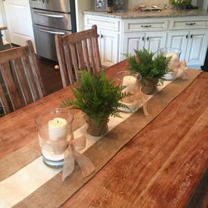 Pode incluir: Uma mesa de madeira com um corredor branco e de juta. H&aacute; tr&ecirc;s casti&ccedil;ais de vidro com velas brancas e la&ccedil;os de juta. Cada casti&ccedil;al &eacute; decorado com uma planta de samambaia em vaso.