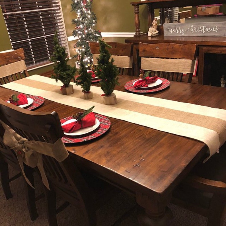 May include: A wooden dining table set for a Christmas dinner with a burlap table runner, red plaid chargers, red napkins, and small Christmas trees as centerpieces. The sign on the back of the table reads "merry christmas".